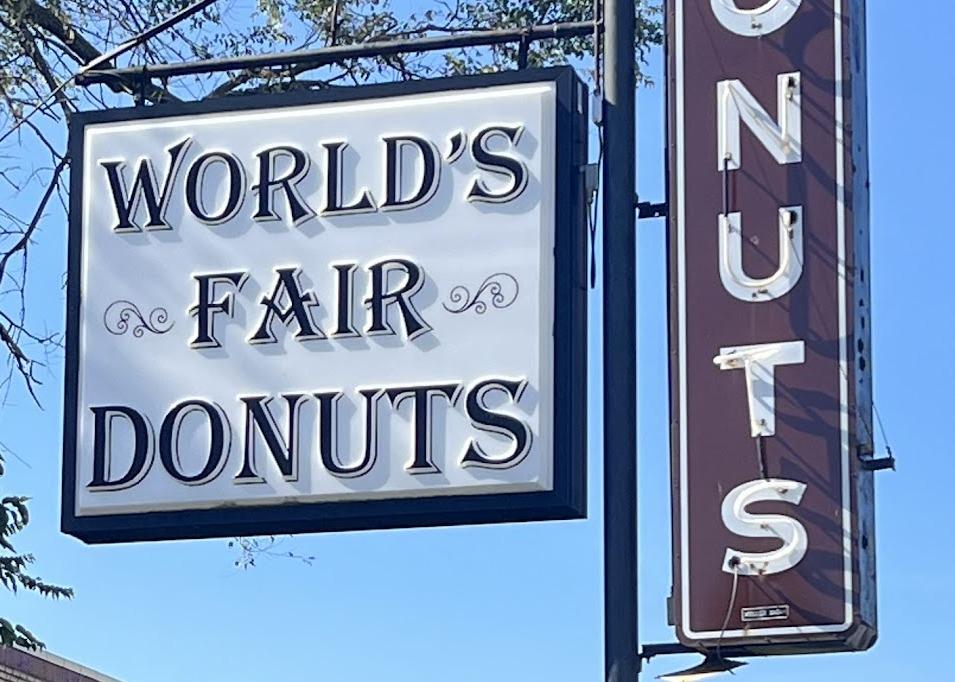 The World's Fair Donuts road sign, a white panel in a dark frame beside a tall oxblood blade sign spelling DONUTS in white neon letters